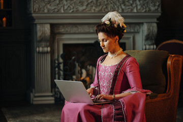 Caucasian middle aged woman wearing Baroque style dress sitting in ornate room using laptop...