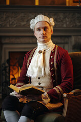 Portrait of young adult Caucasian man wearing historical Baroque costume sitting on chair holding...