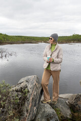 Woman in a beige puffer jacket and gray beanie stands by an Arctic lake holding a turquoise...