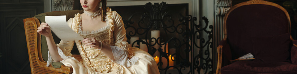 Caucasian young adult woman sitting on ornate chair reading letter in Baroque or Renaissance style...