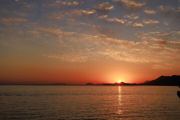 Sun setting behind distant hills, casting golden light across the sea. Wide panoramic view with...