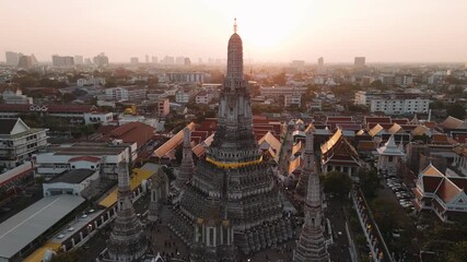 Aerial view of golden wat arun temple, Bangkok, Thailand, against vivid sunset, revealing intricate architectural details near chao phraya river.