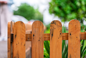 Close up view of a weathered light brown wooden picket fence with slightly rounded tops. Water droplets are visible on the wood, suggesting recent rain. Green foliage is blurred in the background