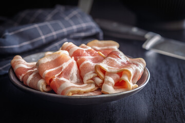 Slices of raw bacon on plate on black table.