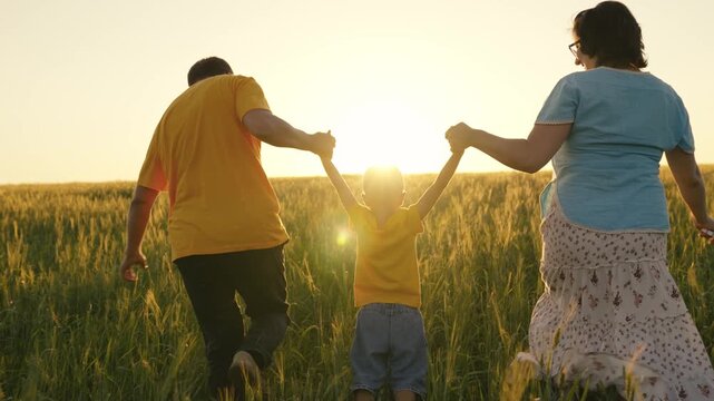 mother and father lead the little kid by the hands along the foam field, concept of family business life, people walk and play with boy son at sunset, fun childhood in nature, enjoy glare of sunlight.