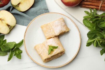 Pieces of tasty apple strudel with powdered sugar, mint, fruits and cinnamon on white marble table,...