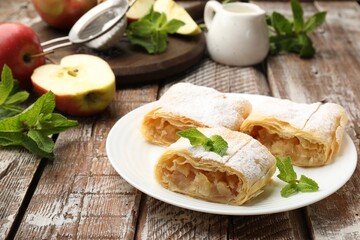 Pieces of tasty apple strudel with powdered sugar, mint and fruits on wooden table, closeup
