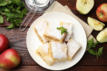 Pieces of tasty apple strudel with powdered sugar, mint and fruits on wooden table, flat lay