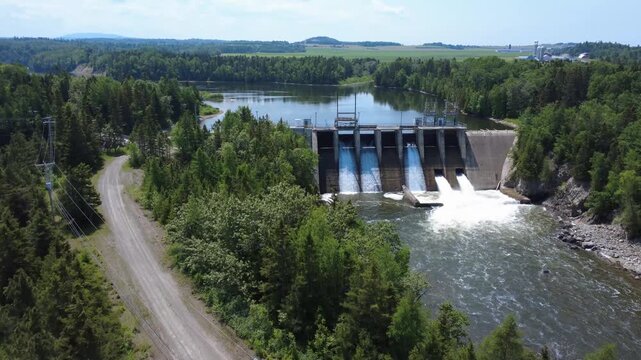 A view of a hydroelectric dam with cascading water and full-flow spillway gates, combining industrial design with a lush forest setting, showcases sustainable renewable energy production. Mitis-2 Dam.