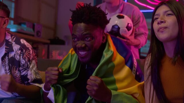 Front view shot of emotional young Black man holding Brazil flag while watching sports match on TV with group of friends in neon lit room