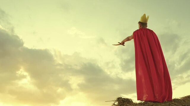 teenage boy red cloak and golden crown his head. child kid plays superhero with sword. child's dream warrior. sky background. happy teenager in halloween costume.. holiday mood superhero child son