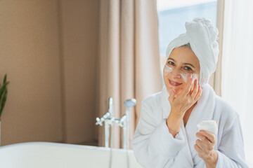 Mature adult old woman applying nourishing anti age face cream doing daily beauty routine. Senior...