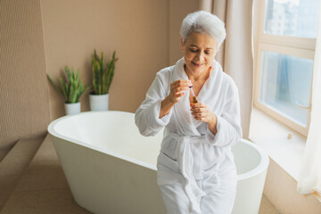 Woman applying natural drops cosmetics oil to face skin. Mature adult old woman applying nourishing...