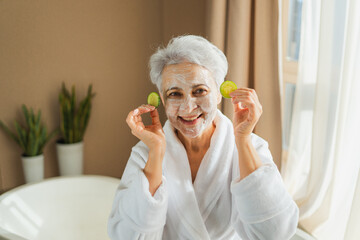 Mature adult old woman with nourishing anti age face cream mask holding cucumber slices. Senior...