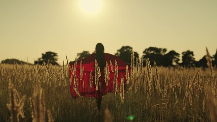 A girl runs across the field in a red superhero cloak waving in the wind at sunset, childhood dream...