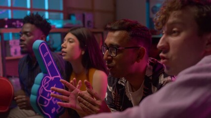 Side view shot of young MIddle Eastern man watching sports match on TV with multiethnic group of friends and jumping up cheering for goal
