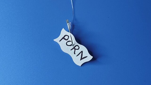 The word porn written on a white paper note and hooked on a fishing hook like bait. Isolated against a blue background with copy space. Stop porn addiction or masturbation habit concept.