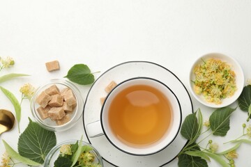 Tasty linden tea in cup, spoon, sugar cubes, leaves and flowers on white table, flat lay