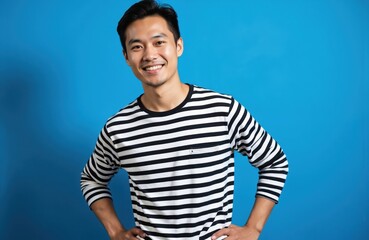 Obraz premium Young Asian man in black and white striped shirt poses with hands on hips. He wears black pants against a clean blue studio background. Confident expression, relaxed posture, bright studio lighting.