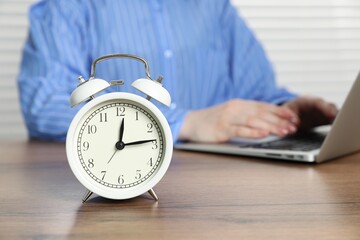 Deadline. Woman with laptop working at wooden table in office, focus on alarm clock