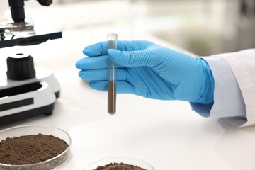 Scientist with test tube of soil sample at white table in laboratory, closeup