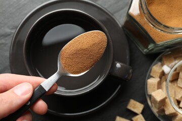 Woman holding spoon with instant coffee above cup at black table, top view