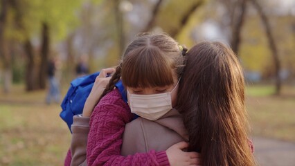 Fototapeta premium mother puts little child a medical mask on her face, protection against coronavirus covid 19, kid with backpack, happy family, modern pandemic life, student health safety, anti corona germs, hug mom.