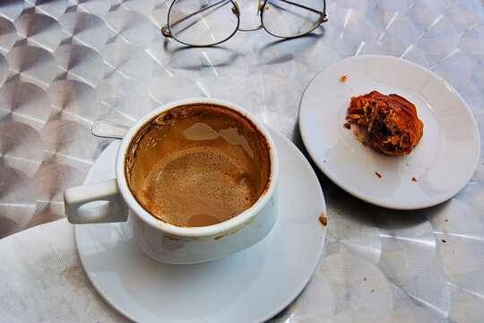 The white ceramic cup with  cappuccino leftovers with creamy foam, last bite of sfogliatella paistry and glasses on a metallic table in outdoor cafe. Selective focus. 
