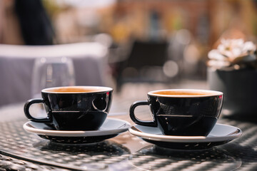 Two dark coffee cups sit on a table in a sunny outdoor cafe setting