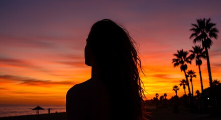 Silhouette of a woman with palm trees during a vibrant sunset on a beach with orange and purple sky