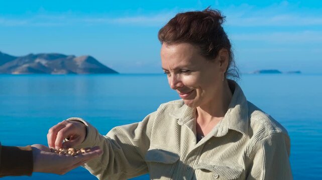 Woman choking on peanuts. Woman takes some peanuts from a person's hand, eats them and immediately starts choking, holding her throat with both hands near a lake and a clear sky
