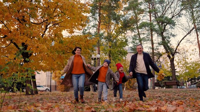 Kids, mom, dad play together, holding hands in autumn outdoors. Happy kids play, run, holding hands of mom dad, along yellow autumn leaves in park. Weekend family walk, mother, child, father in park - Powered by Adobe