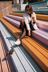 Woman enjoys snack while sitting on colorful steps with a curious pigeon nearby © Irina Magrelo
