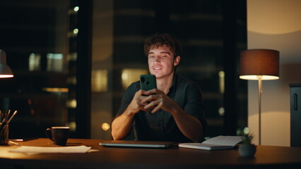 Employee messaging smartphone night office under lamp light. Smiling businessman