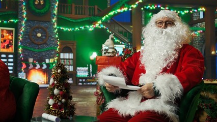 Man dressed as Santa Claus maintaining jolly demeanor reading letters received from kids. Person...