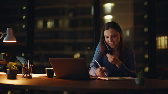 Multitasking lady talking smartphone writing data in notebook at night office.