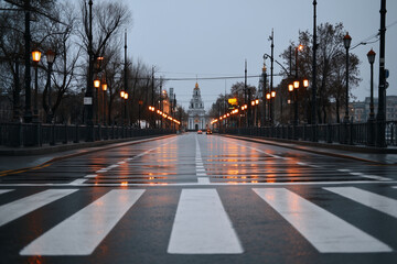 Empty pedestrian crossing on wet city street at dusk, with illuminated streetlights reflecting on the wet asphalt, leading towards prominent building.