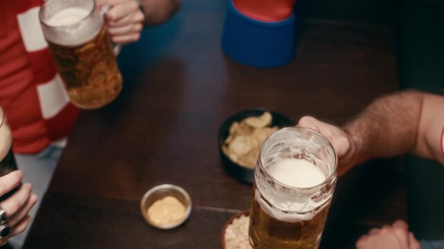 High angle of group of young friends sitting by table in bar after watching football broadcast, clinking with glasses of beer and enjoying foaming beverage