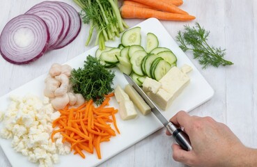Close-up of knife, vegetable slices on cutting board. Fresh vegetables scattered around on white countertop. Carrot, cucumber, green onions, red cabbage, lettuce, cheese. Hands of cook cutting