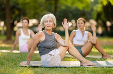 Smiling senior silver-haired woman enjoying group yoga practice outdoors, sitting on mat on green...