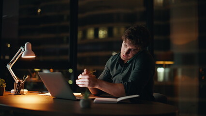 Overloaded freelancer calling cellphone looking laptop at office late evening.