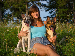 PORTRAIT, CLOSE UP: Woman with a beaming smile embraces her two dogs in a vibrant, sunlit meadow....
