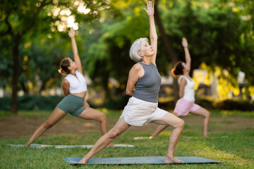 Elderly woman doing warrior yoga pose on mat in group of women in park