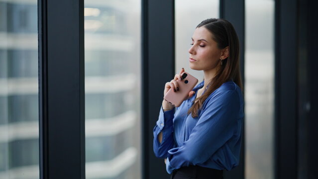 Thoughtful business owner standing by office window holding smartphone closeup. - Powered by Adobe