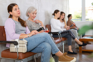 Portrait of bored positive mature woman with phone looking away while waiting in line for job...