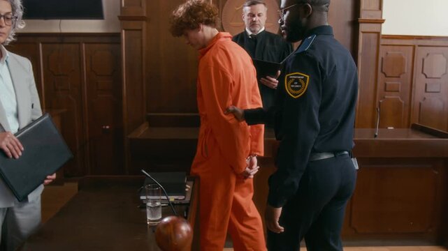 African American security officer bringing young red-haired criminal in prison robe and handcuffs to court to be tried