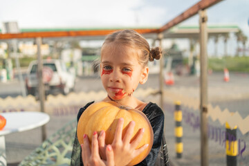 Child dressed in a spooky costume holds a bright orange Halloween pumpkin with joy. The backdrop...