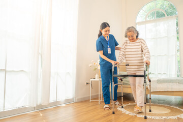 Female caregiver take care and help senior woman using walker walking in living room. Retirement...