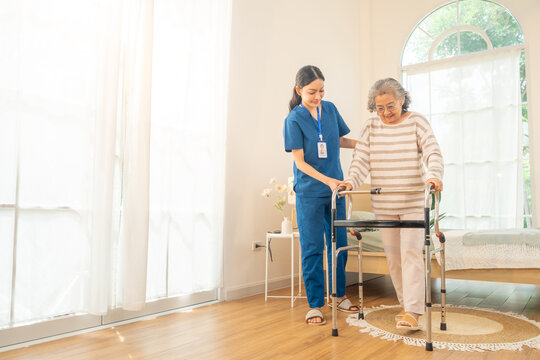 Female caregiver take care and help senior woman using walker walking in living room. Retirement rehabilitation nursing home. Elderly people health, home medical wellness healthcare services concept.