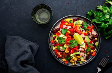 Tex-mex salad with canned tuna, avocado, corn, red beans, cherry tomatoes, cilantro and lime dressing, black table background, top view